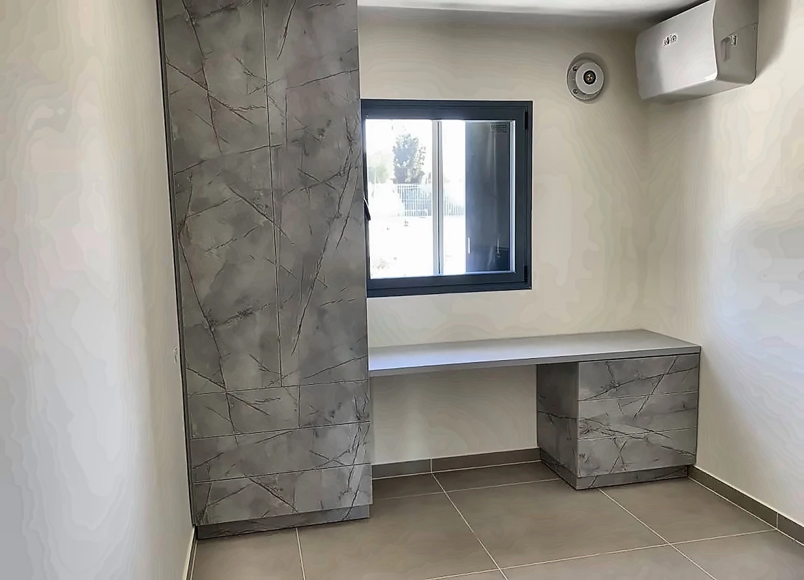 חדר עבודה עם ארון גבוה ושולחן כתיבה עם מגירות בחיפוי דמוי אבן אפורה, מתחת לחלון אלומיניום